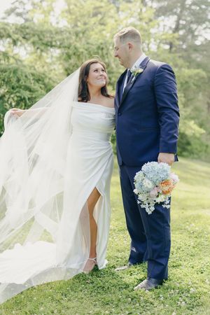 A couple in wedding attire share a romantic moment outdoors, with a flowing white veil and blue hydrangea bouquet.