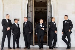 Groomsmen in black tuxedos posing playfully along white columned building.
