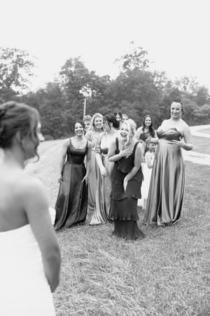 Black and white photo of bridesmaids in flowing dresses reacting with joy during a wedding photoshoot outdoors.