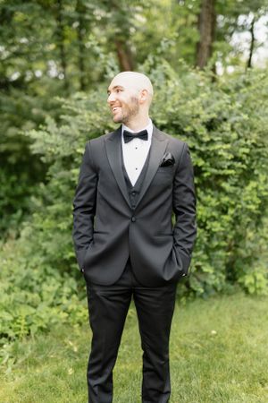 Groom in classic black tuxedo poses in outdoor garden setting.