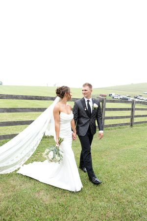 A couple in wedding attire poses together in a grassy field with wooden fencing in the background.