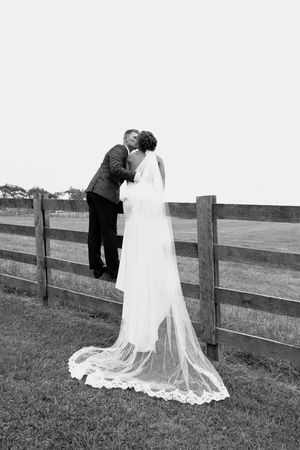 A romantic black and white wedding photo showing a couple by a wooden fence with the bride's long lace veil trailing behind.