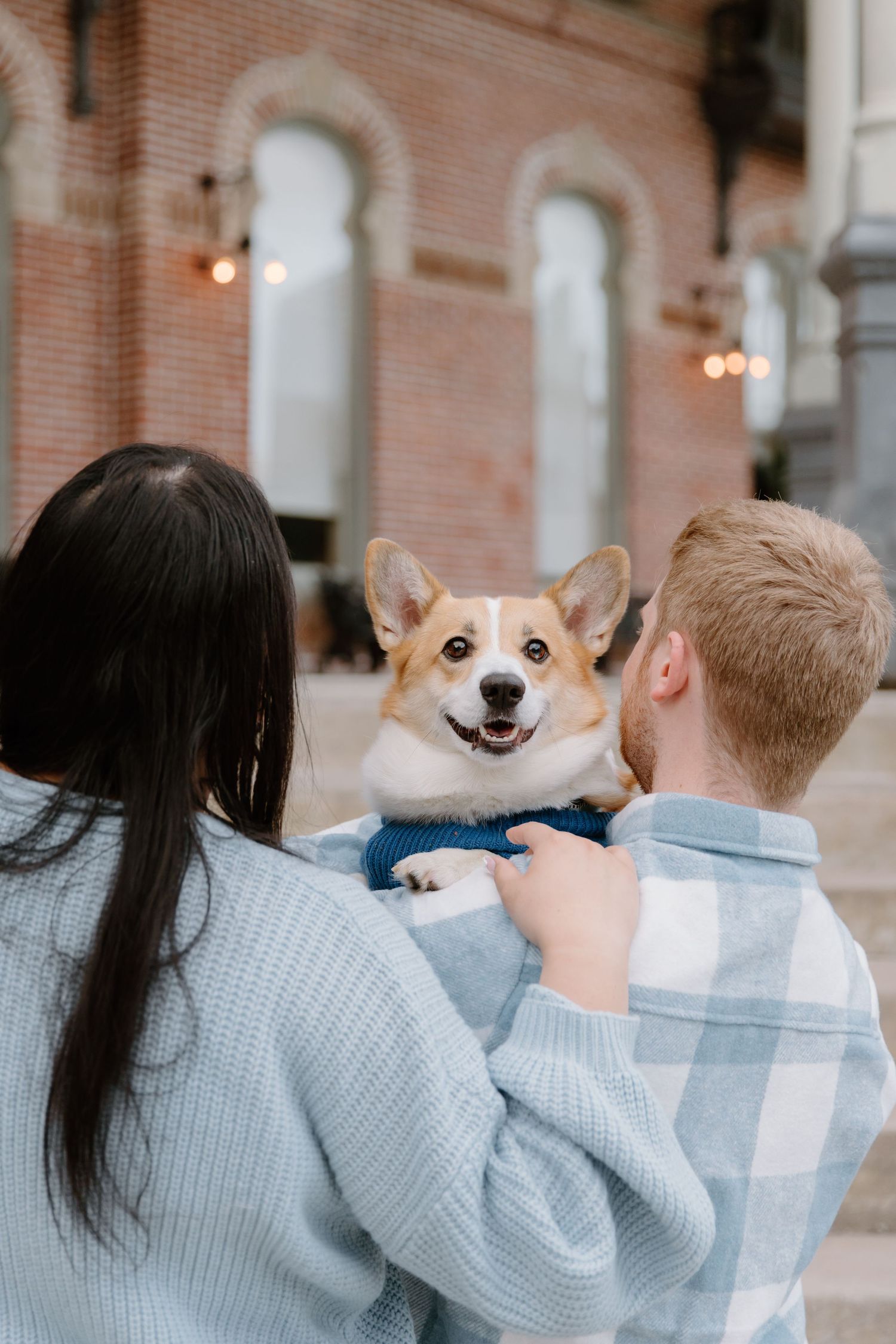 Happy Corgi dog smiles while being held by couple in casual outfits near brick building.