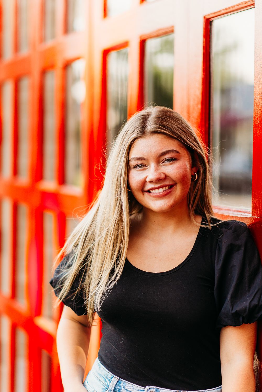 Woman in black top poses in front of vibrant red windows with a cheerful expression.