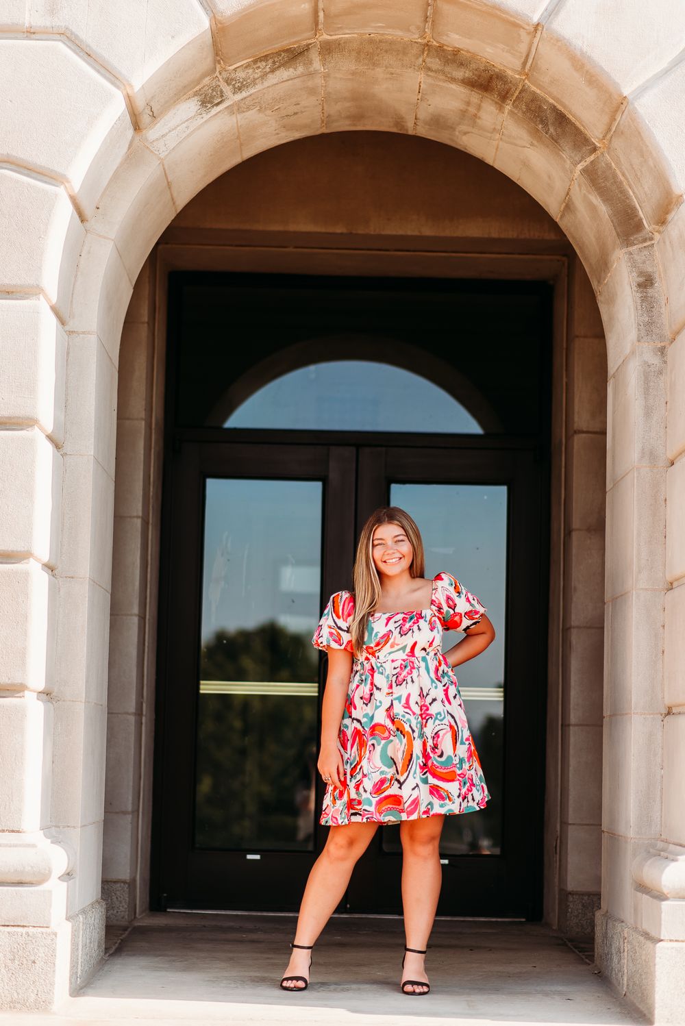 Woman in floral print dress poses confidently under stone archway entrance.