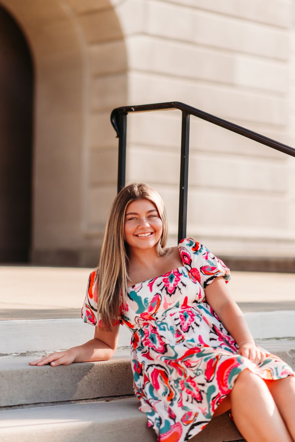 Woman in floral dress sits on stone steps with joyful expression.