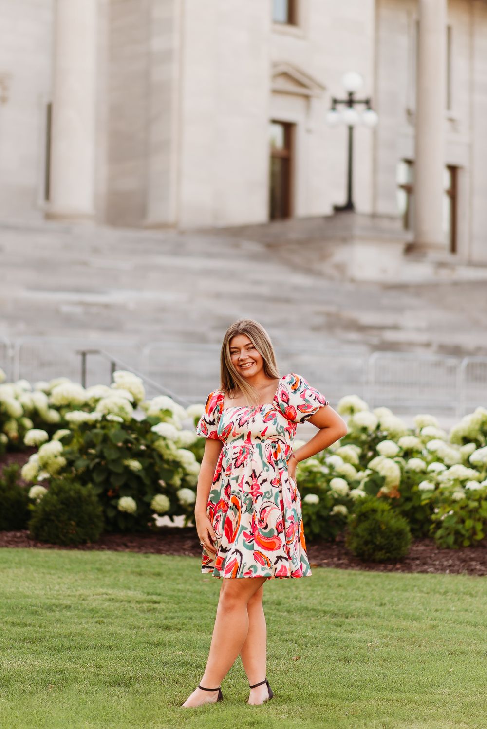 Woman in a red and white floral dress poses on manicured lawn near architectural columns.