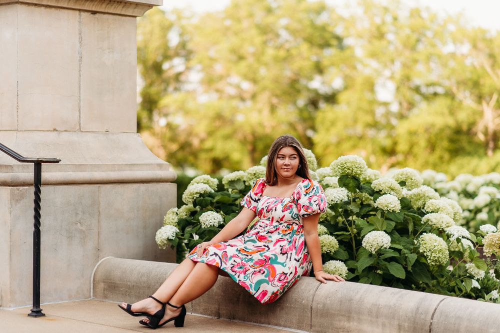 Person in floral dress sits casually beside blooming hydrangea bushes near a classical building.