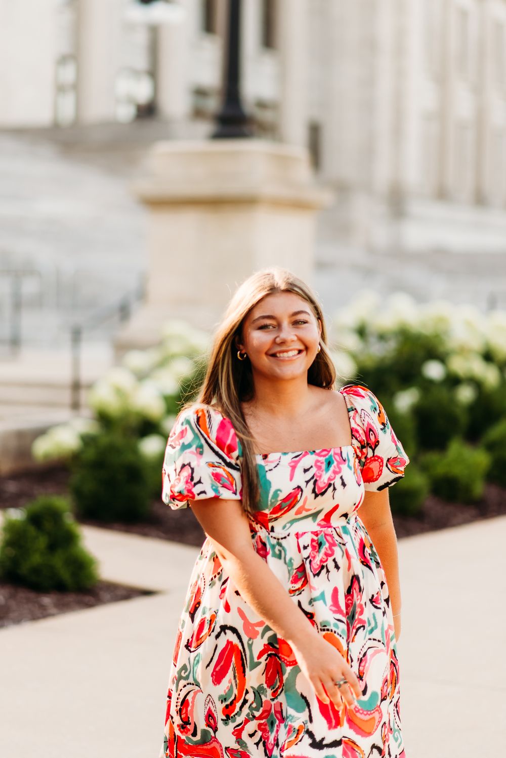 Woman wearing a colorful floral dress smiles brightly while standing near a stone building.