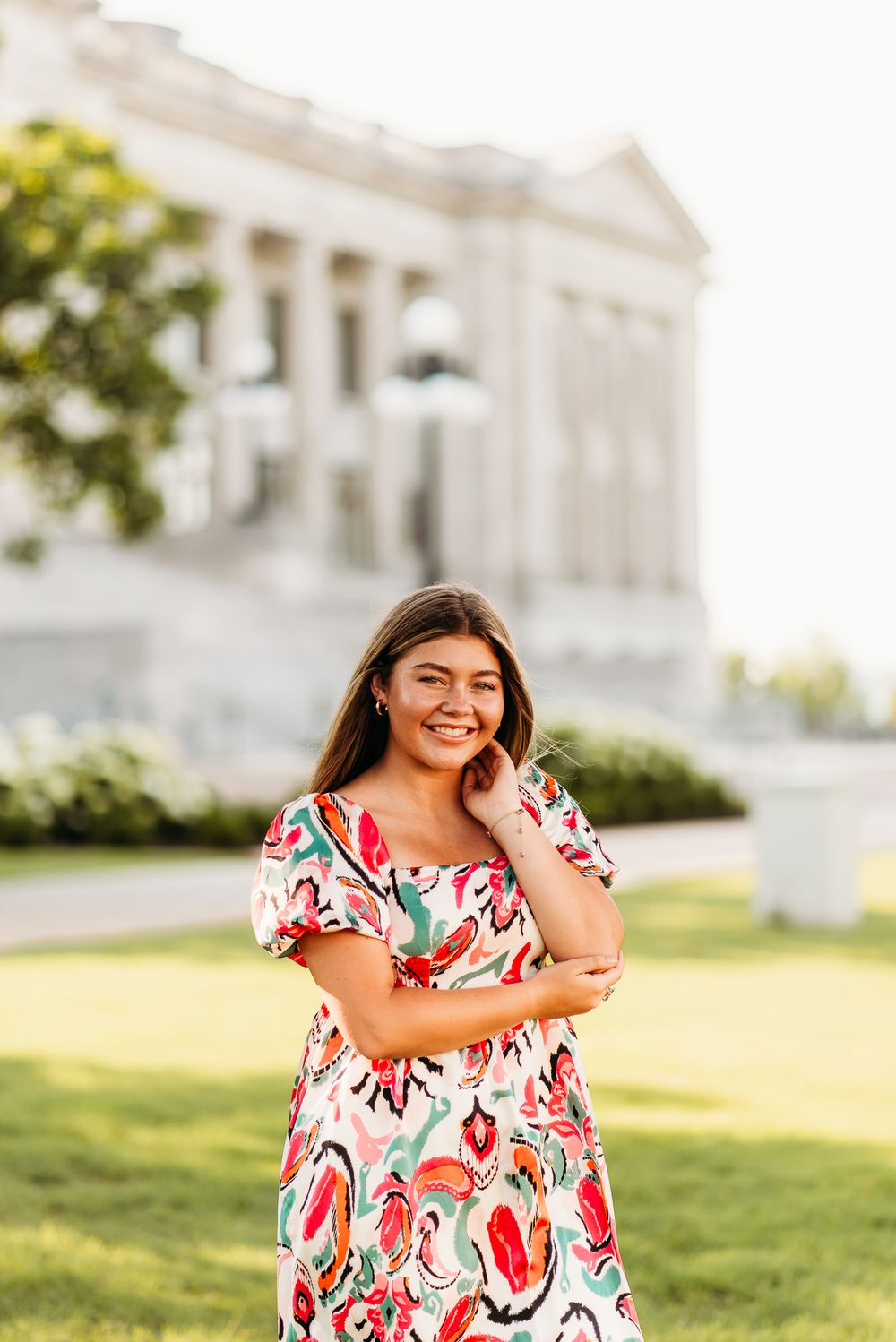 A young woman in a floral dress poses outdoors in front of a classical building on a sunny day.