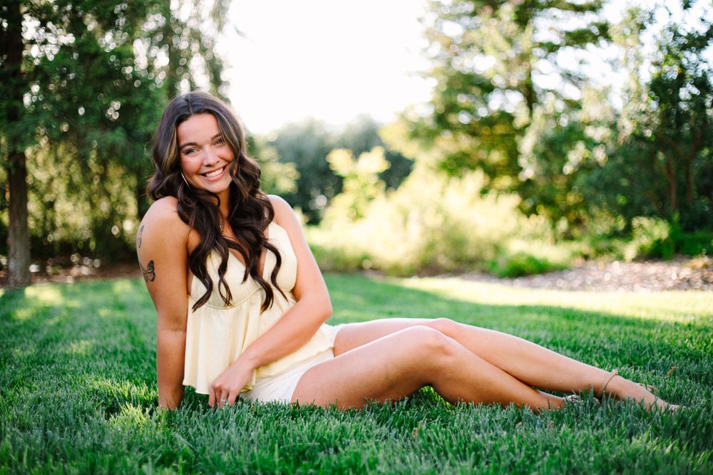 Woman in cream-colored dress sitting on lush green grass at sunset in a park setting.