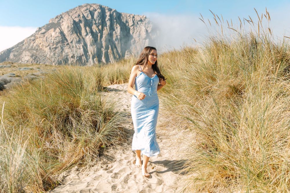 Someone in a blue dress walks along a sandy beach path with tall grass and a mountain in the background.