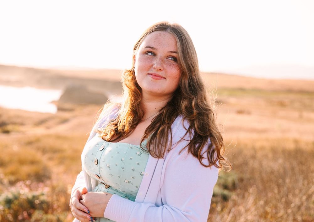 Woman in white sweater holding a green mug and smiling while standing in golden field at sunset.