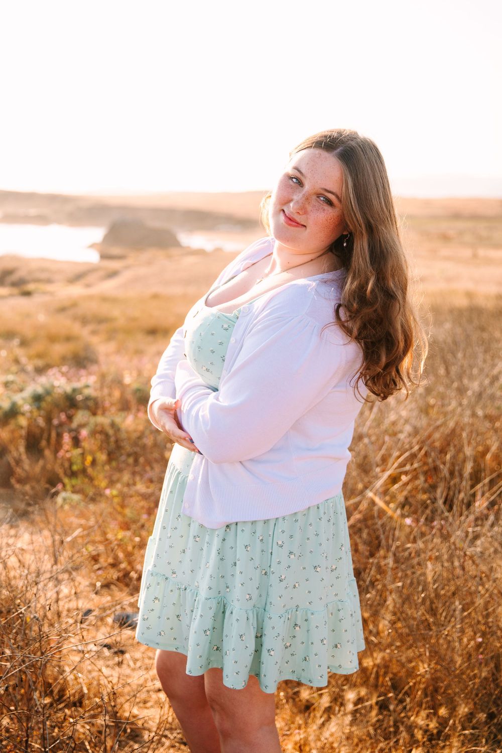 Four dreamy sunset photos show someone wearing a white top and mint floral skirt posing in golden grass at magic hour.