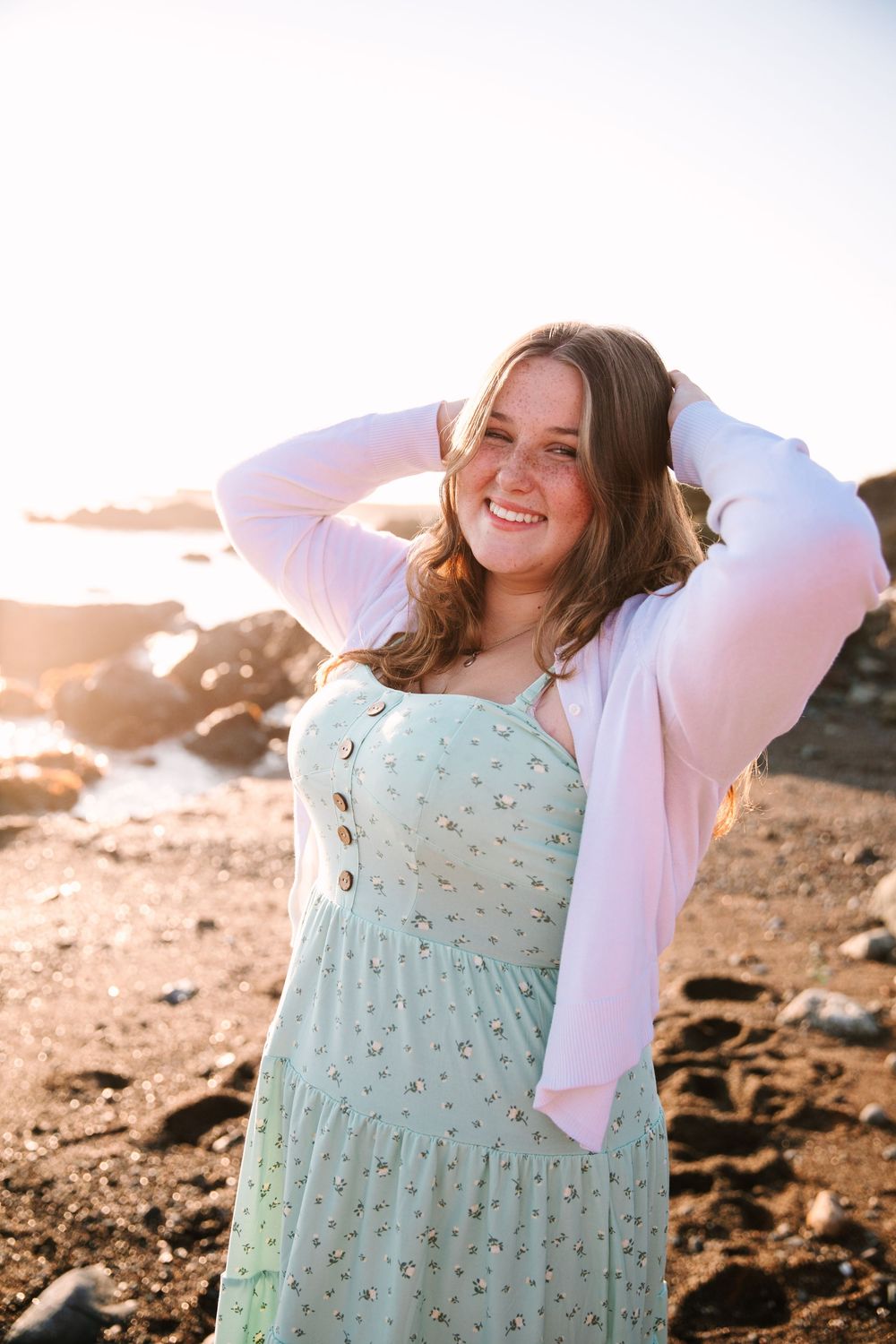 A person in a mint green dress poses against a rocky coastal backdrop during golden hour sunset lighting.