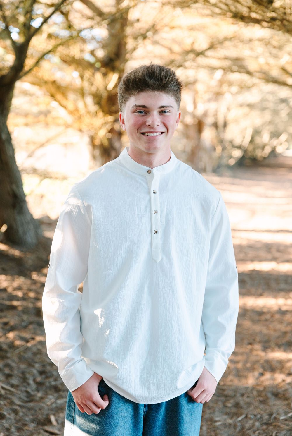 Young person wearing a white henley shirt standing on a forest path in warm autumn lighting.