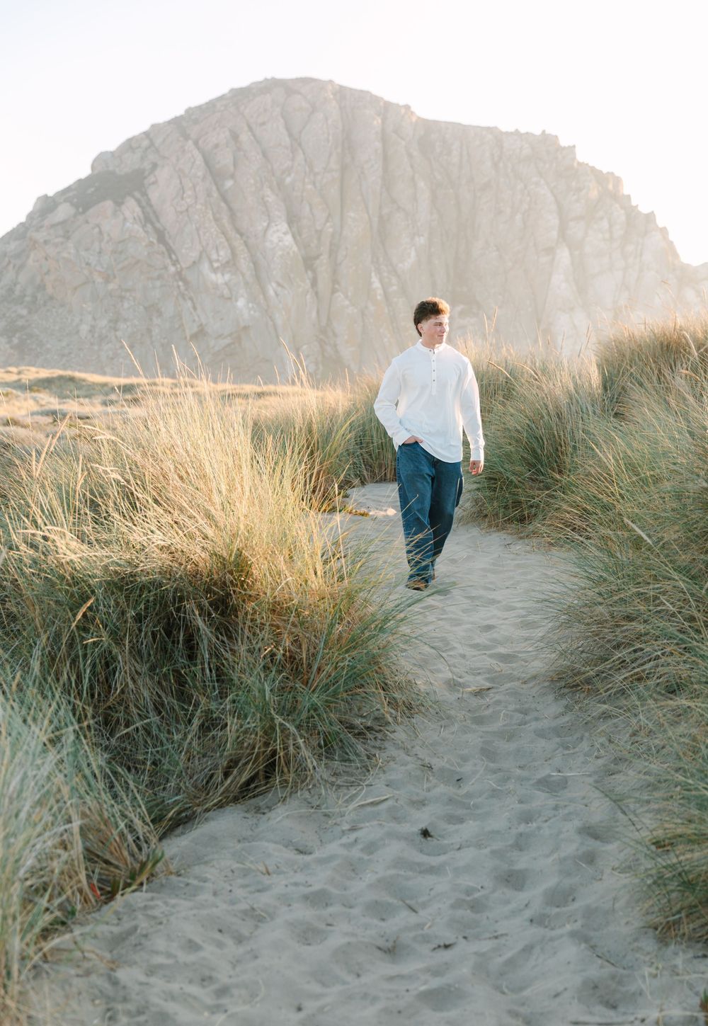 Person walking on sandy path through beach grass at dusk with mountains in background.