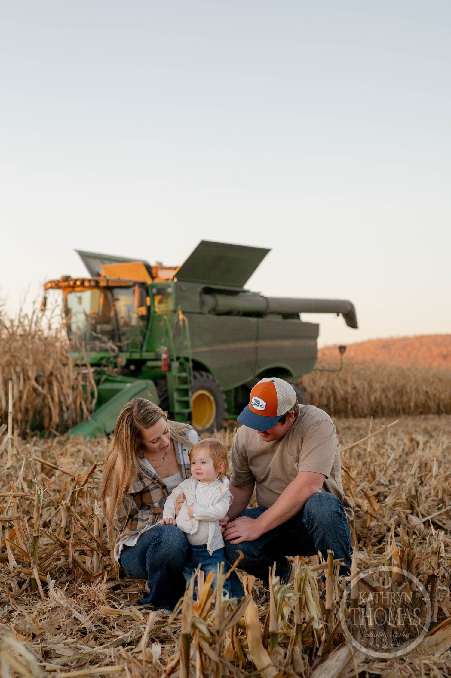 Family gathers in harvested cornfield during sunset with farm equipment in background.