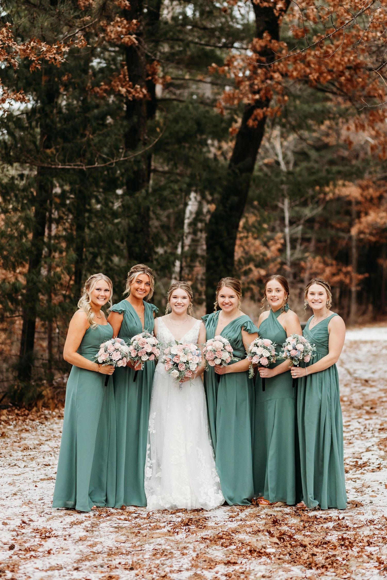 Bridesmaids in matching sage green dresses pose with bride in white gown holding bouquets in autumn forest setting.