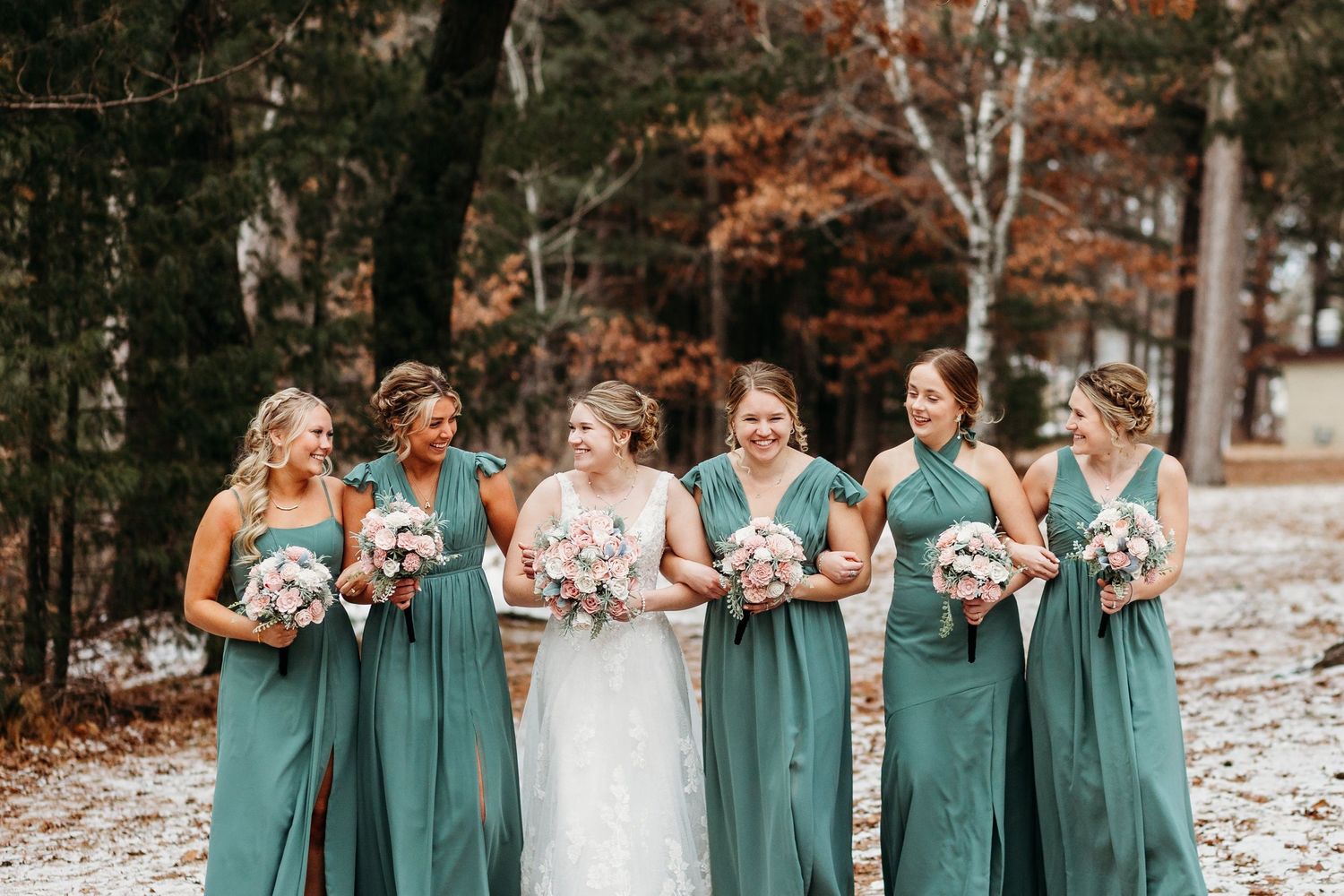 A wedding party wearing sage green bridesmaid dresses and white hold bouquets in a fall forest setting.