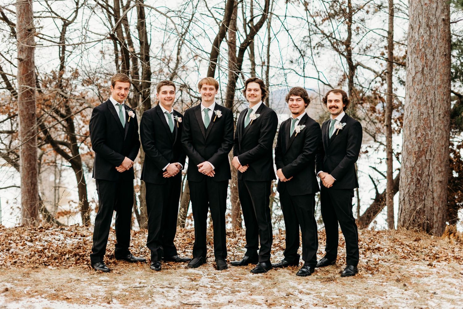 A group of six people stand together wearing matching black suits and ties in an autumn forest setting.