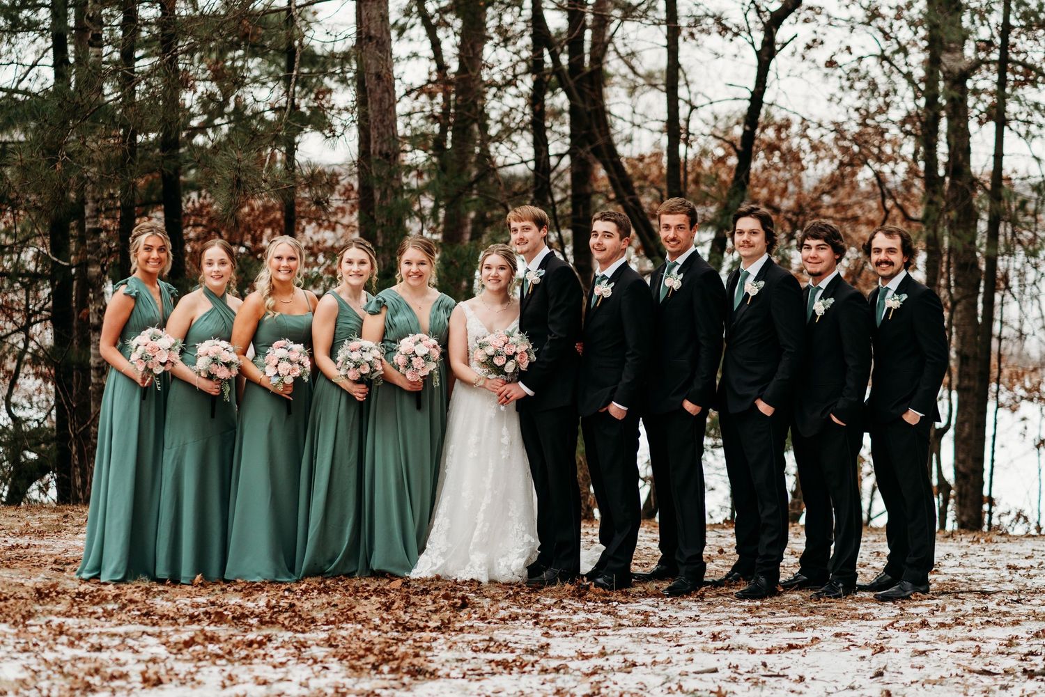 Wedding party in formal attire poses outdoors with bridesmaids in sage green dresses and groomsmen in black suits on autumn day.