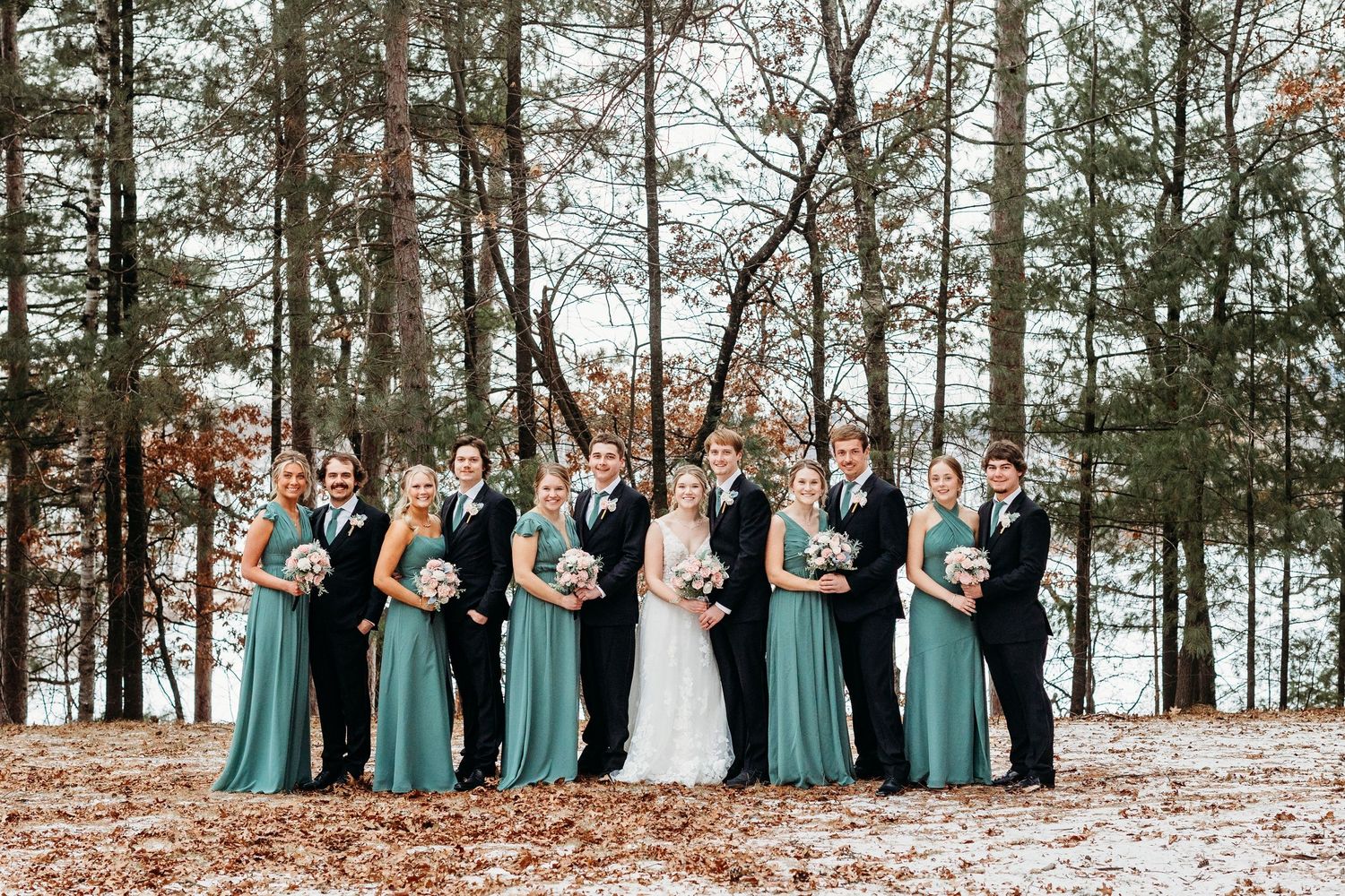 Wedding party in teal bridesmaid dresses poses together on fallen autumn leaves in a pine forest setting.