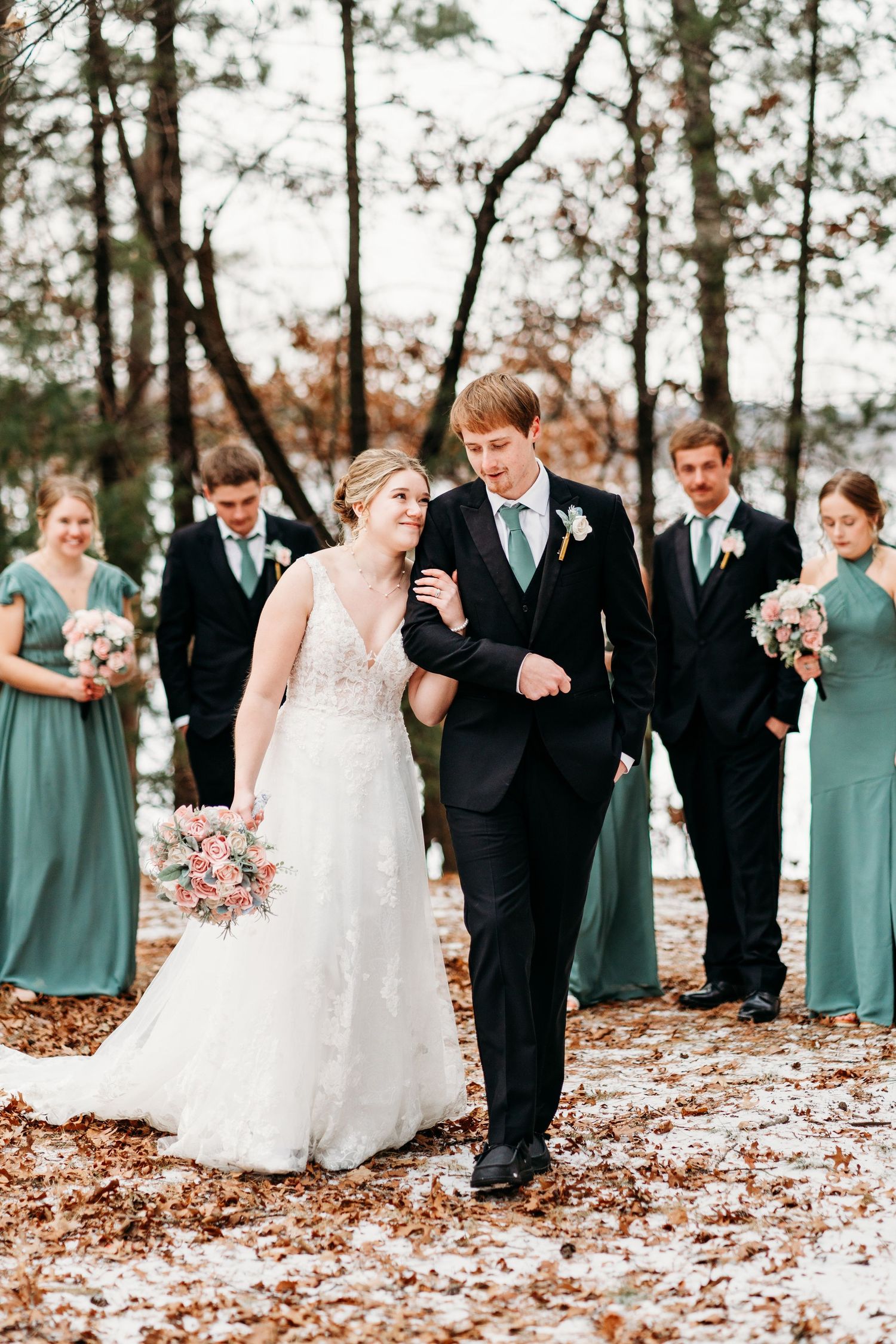 Wedding couple walking through fallen leaves in autumn forest with bridal party in teal dresses in background.