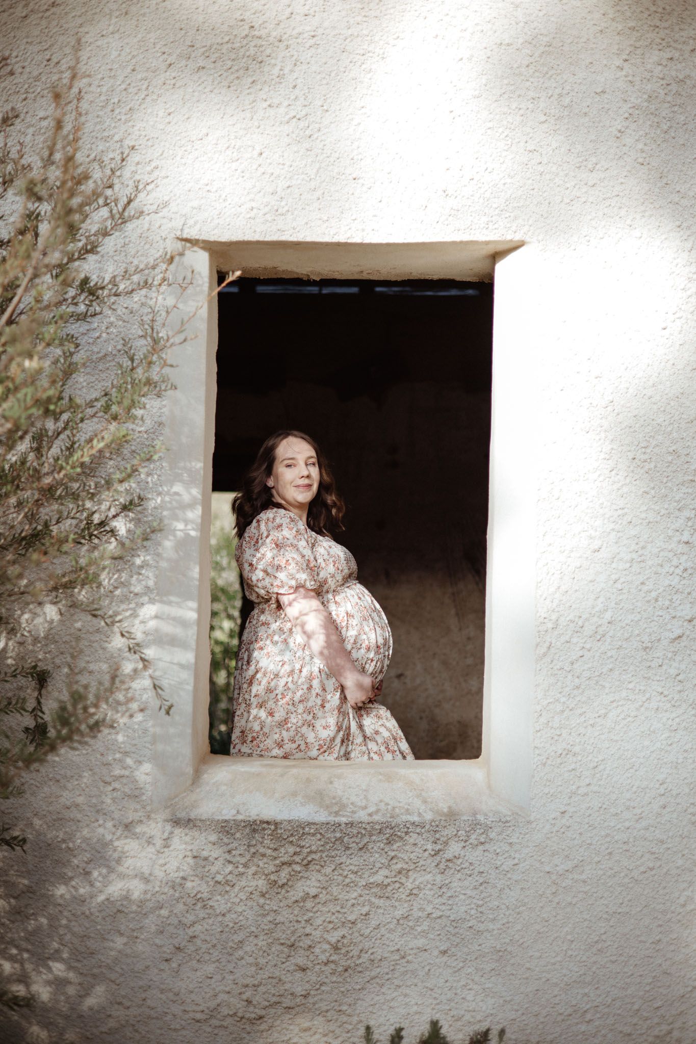 Figure in a floral dress poses in a white stone window frame during a maternity photoshoot.
