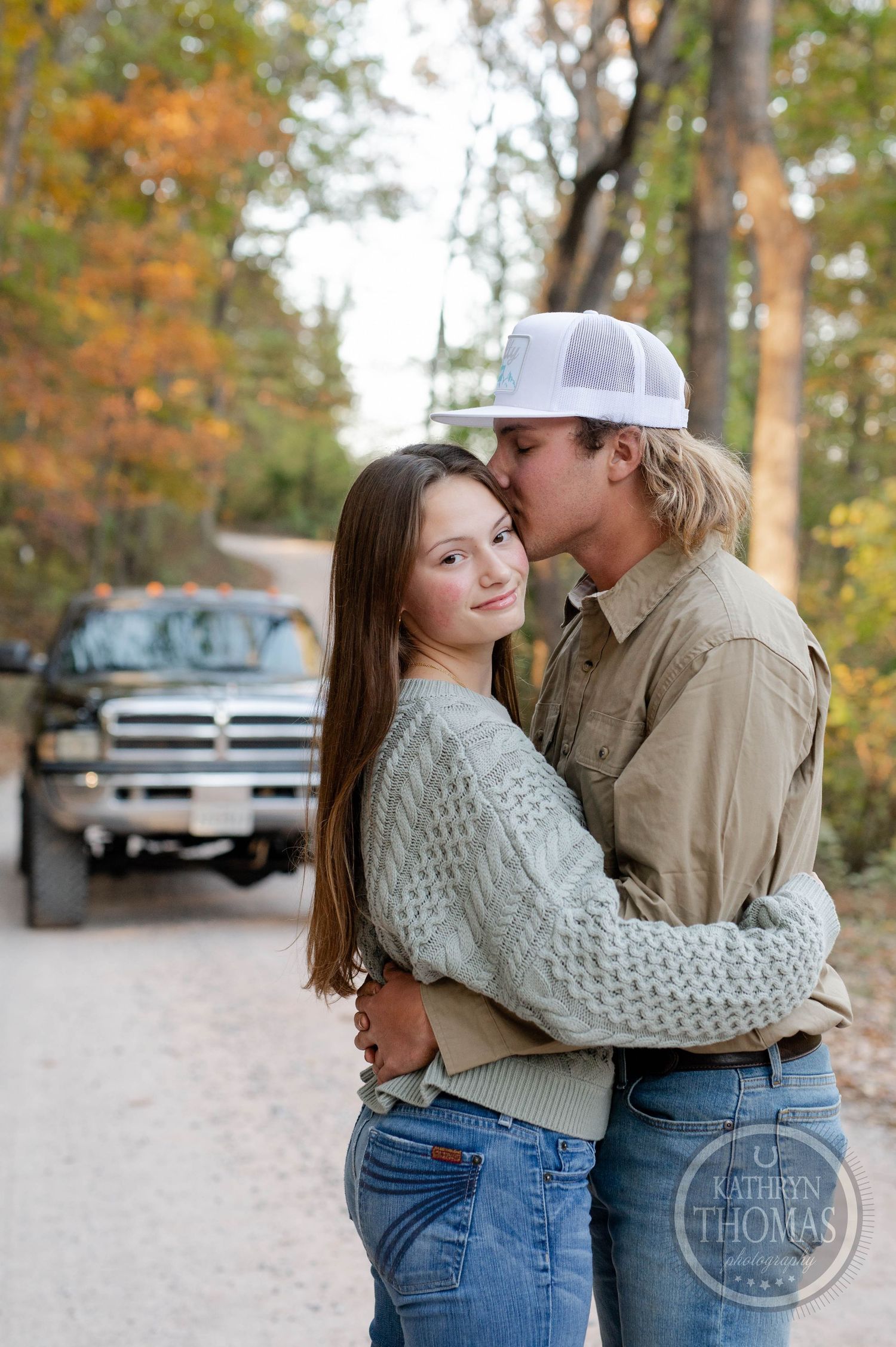 A couple in casual attire embraces on a dirt road surrounded by autumn trees.