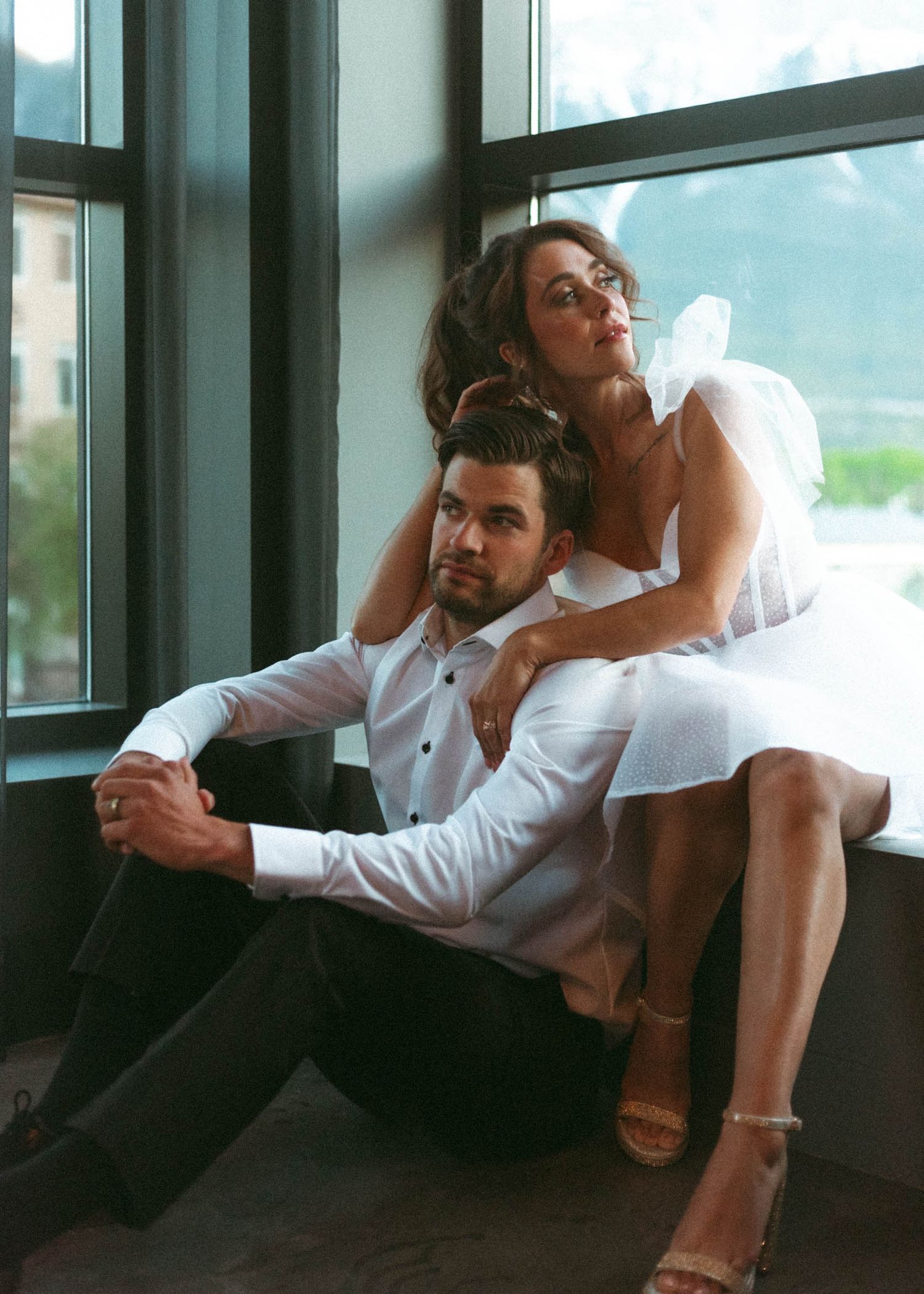 A romantic couple in white outfits pose together near large windows in an intimate fashion photoshoot setting.
