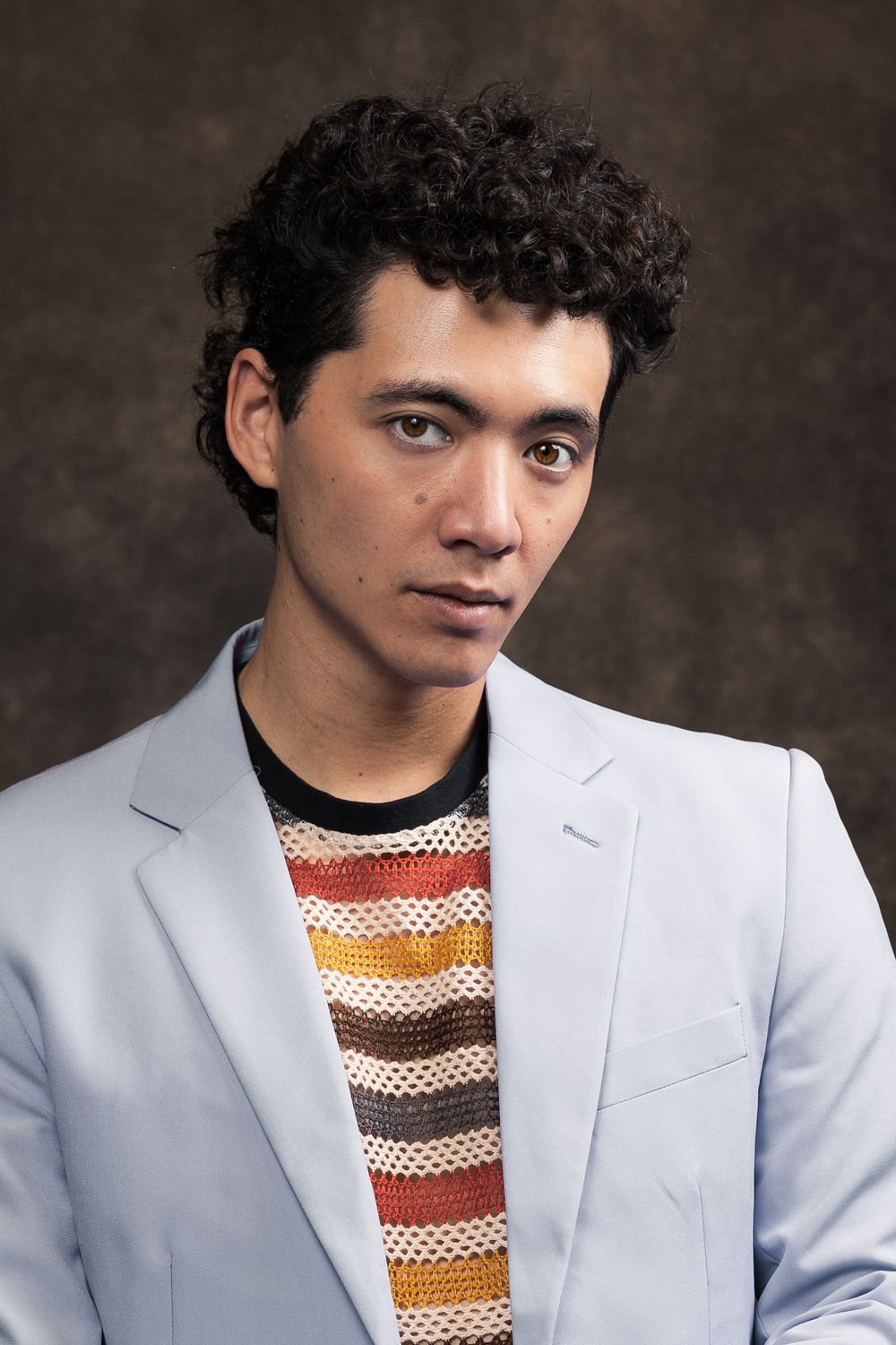 A professional portrait of a young man with curly hair wearing a light blue blazer and striped knit shirt against dark background.