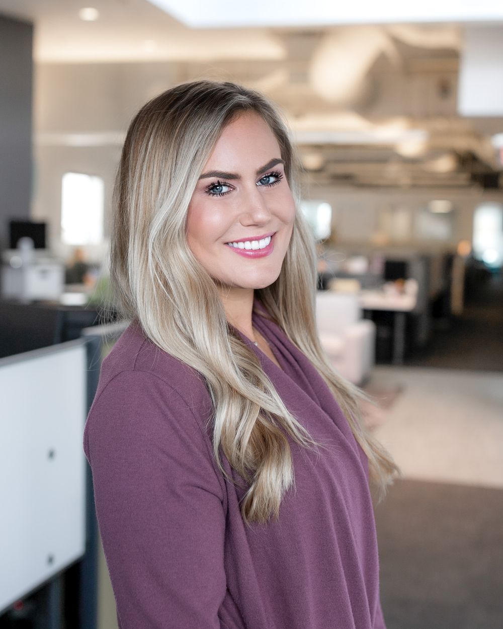 A professional headshot of a smiling blonde woman in a purple top poses in a modern open-plan office environment.
