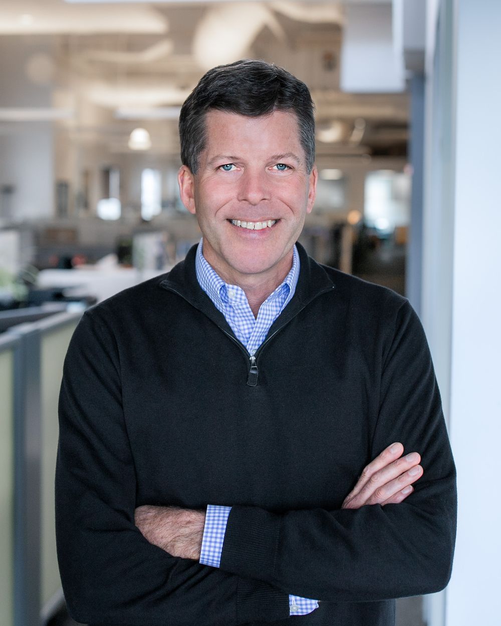 A professional headshot of a middle-aged man in black sweater with arms crossed in a bright office setting.