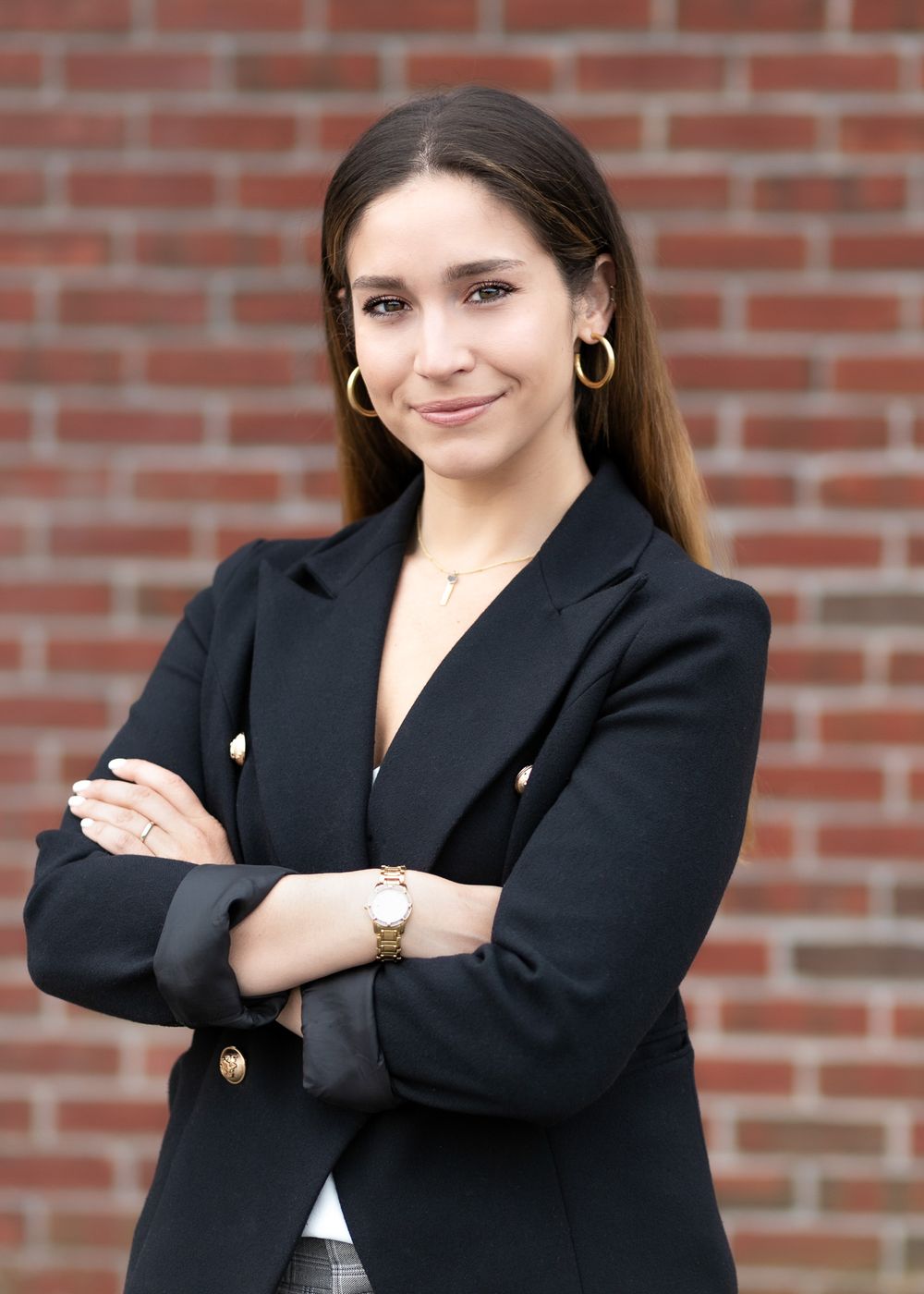 Professional woman in black blazer with arms crossed poses against a brick wall background.