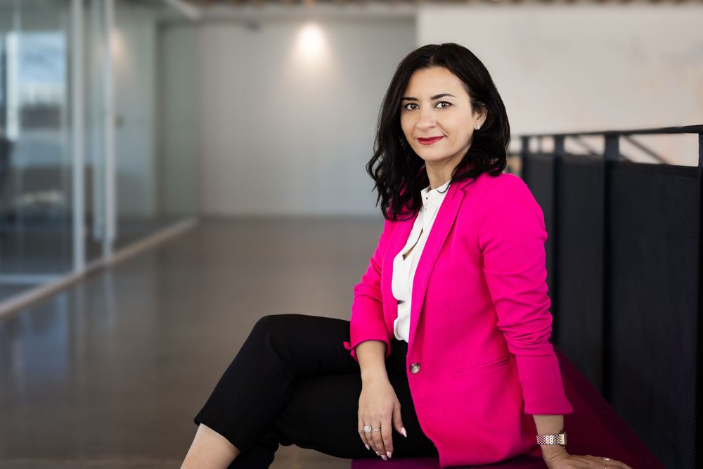 Confident businesswoman in pink blazer sitting in modern office space with bright natural lighting.