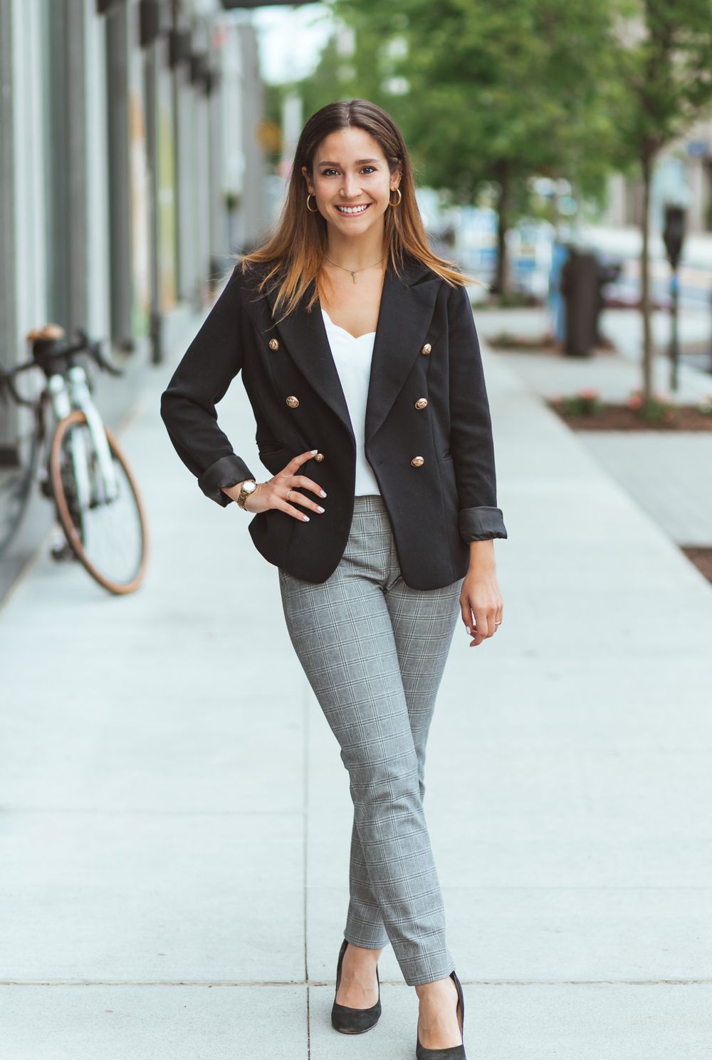 Smiling professional woman in black blazer and gray pants posing on urban sidewalk near bicycle.