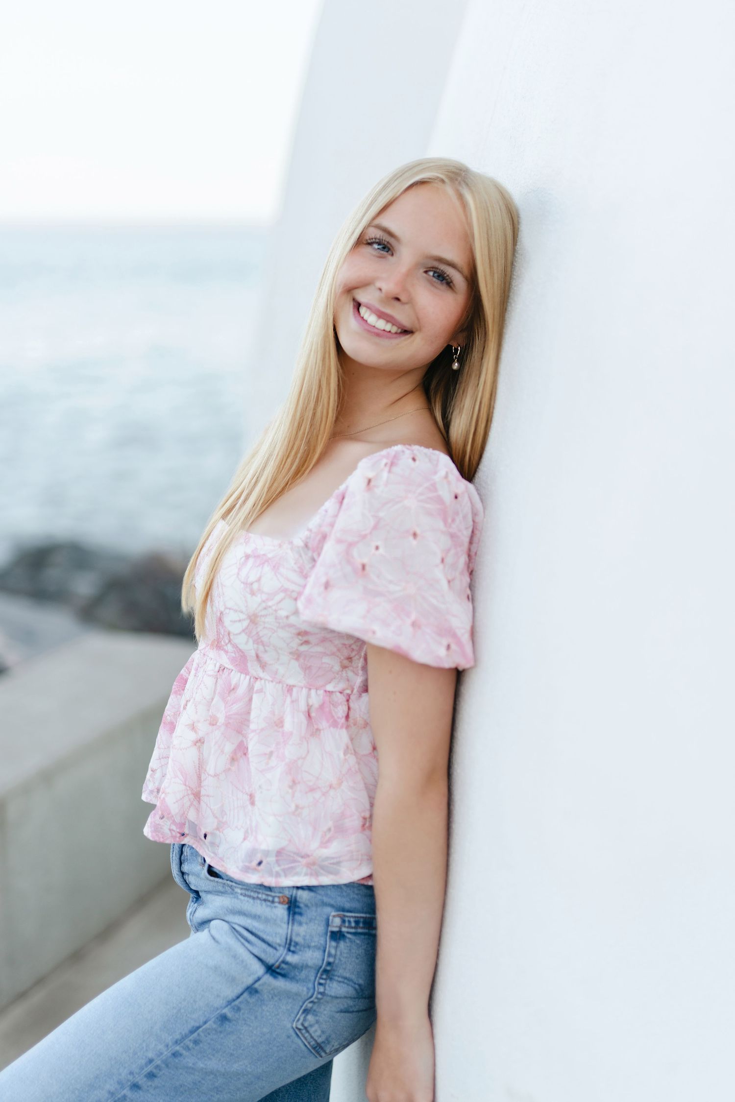 Young woman in black tank top and jeans stands by red railing at waterfront on overcast day.