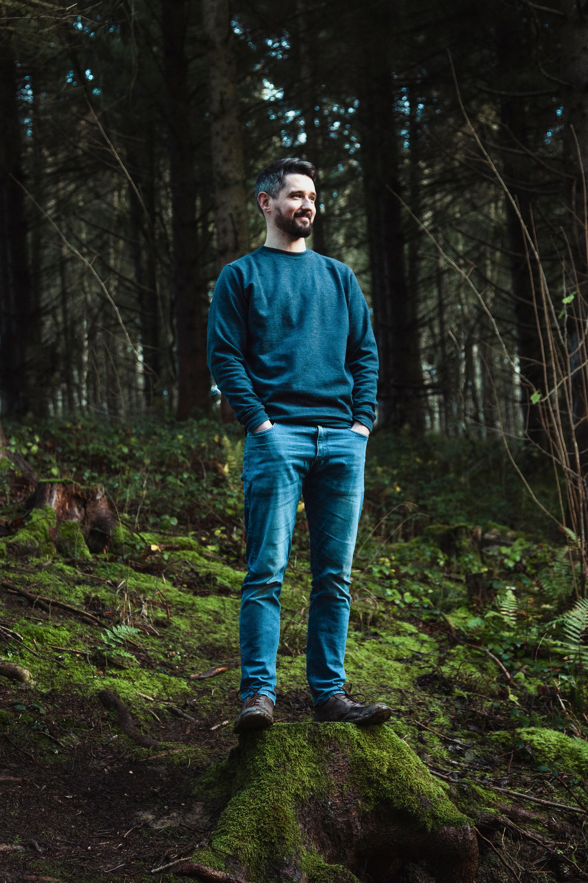 Person wearing blue denim stands on moss-covered ground in a dark evergreen forest.