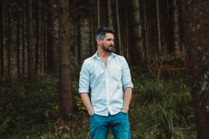 Figure in light blue shirt stands among tall trees in a moody forest setting.