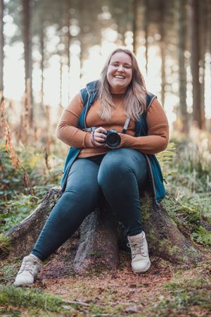 Person in brown jacket and jeans sits on tree stump in sunlit forest.