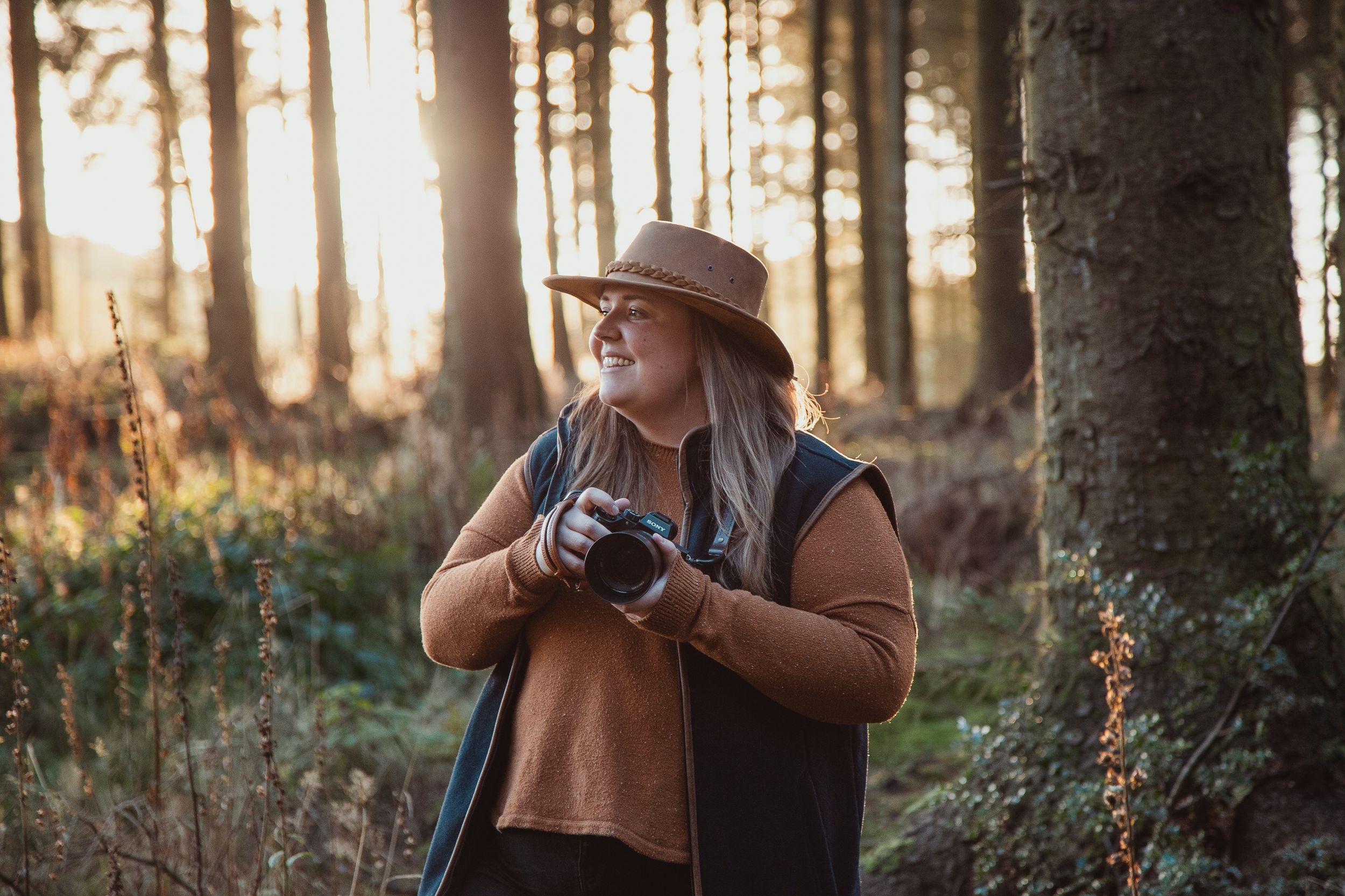 Photographer with camera explores sunny forest wearing brown jacket.