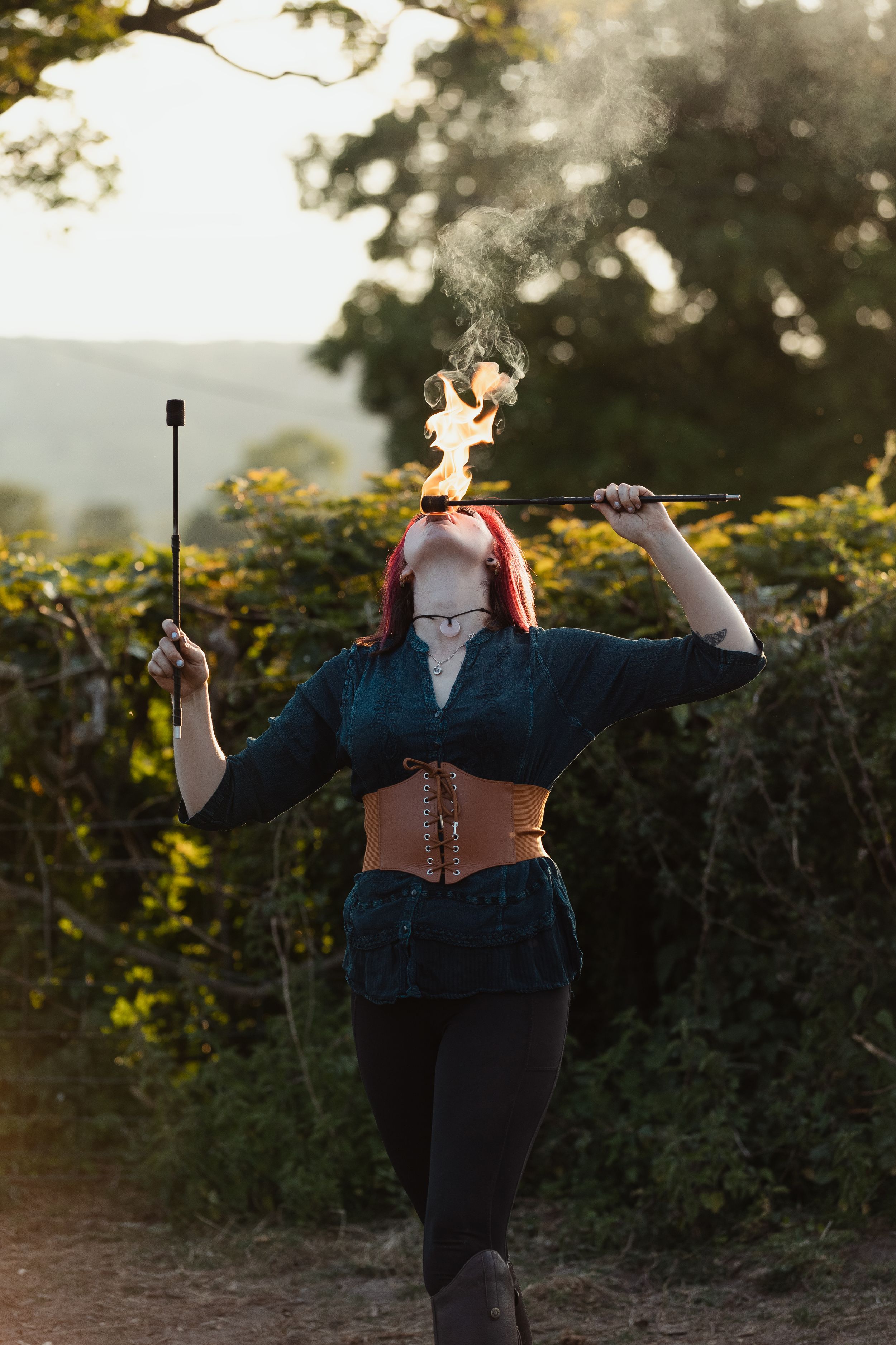 Fire dancer in dark clothing performing with flaming props at dusk.