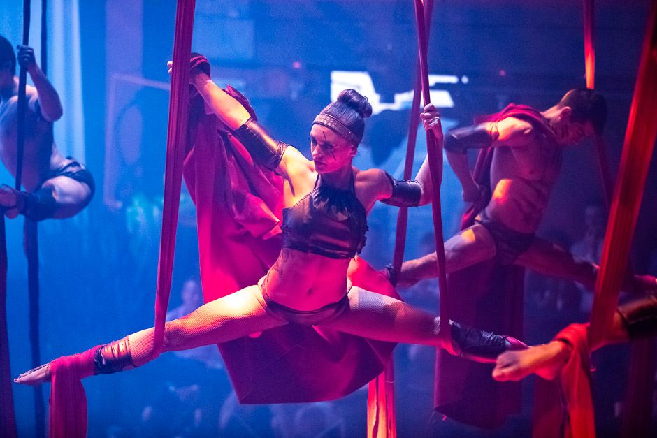 Aerial performer in red costume splits dramatically between silk ribbons in blue-lit theatrical performance space.