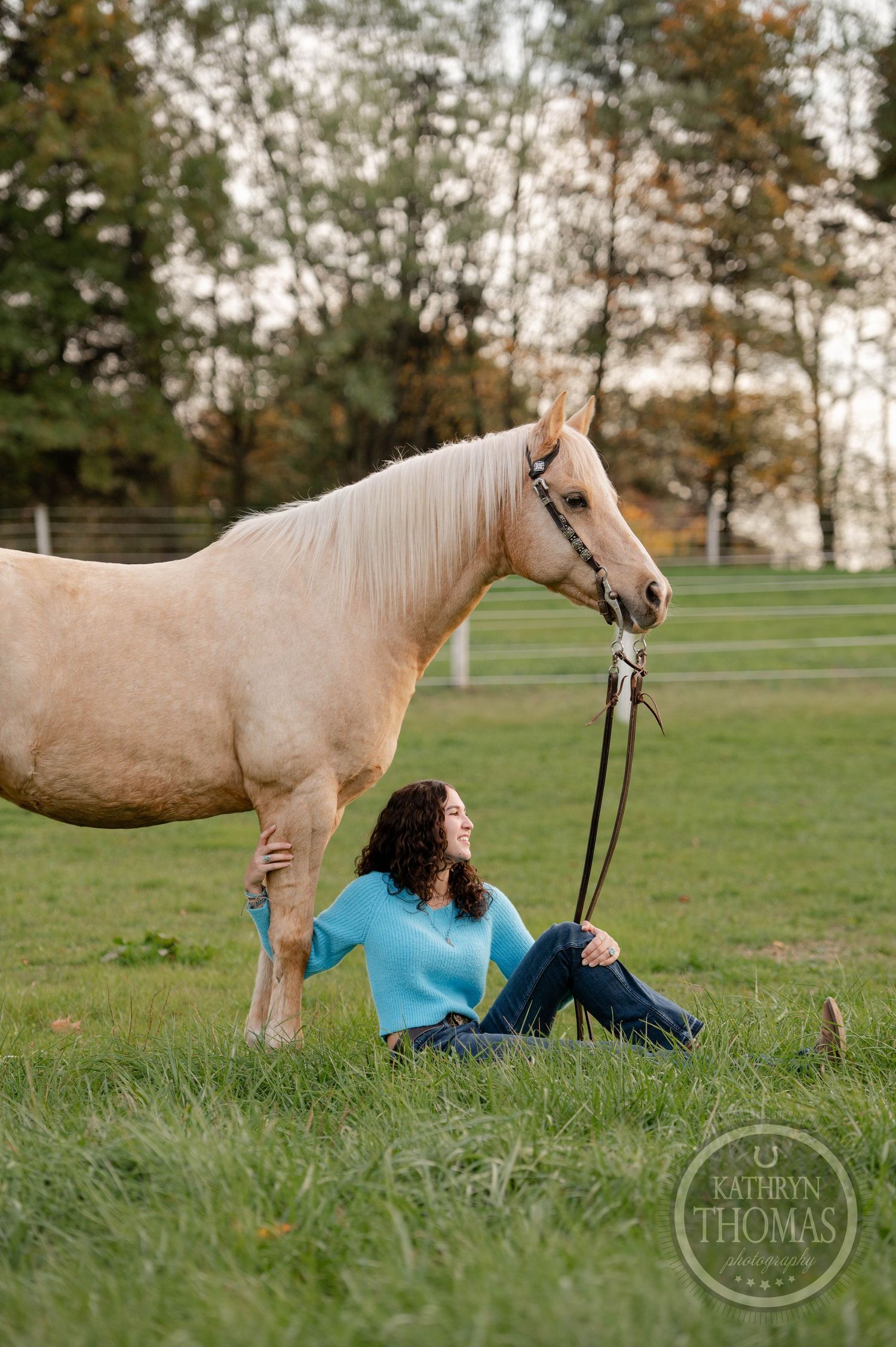 A palomino horse stands beside someone in blue sitting on autumn grass in a fenced pasture.