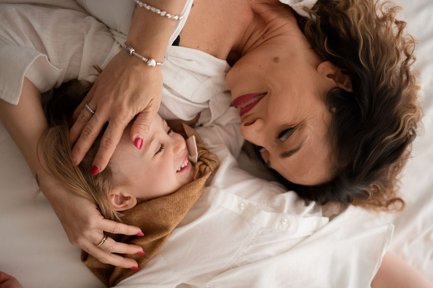 Mother tenderly cradling her young blonde daughter's head while lying together on white bedding.