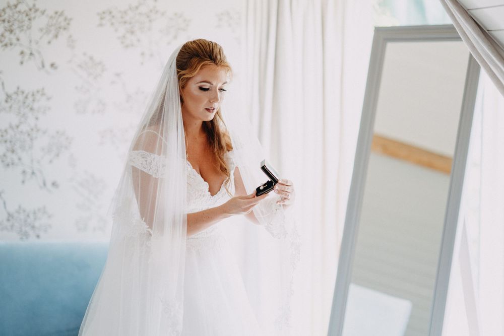 Bride in white wedding gown and veil applies makeup while getting ready, South Farm Cambridgeshire.