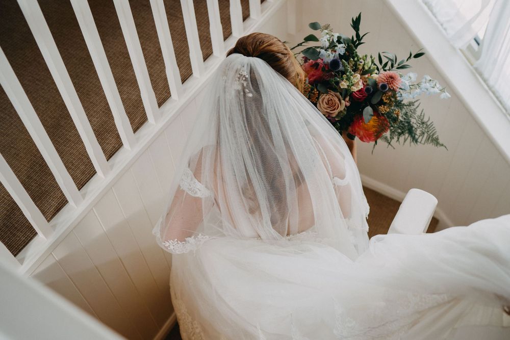 A bride in a white wedding dress and veil sits on stairs next to a colorful floral arrangement.
