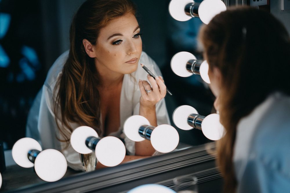 Woman applies makeup in front of a vanity mirror with round bulbs creating a glamorous atmosphere.