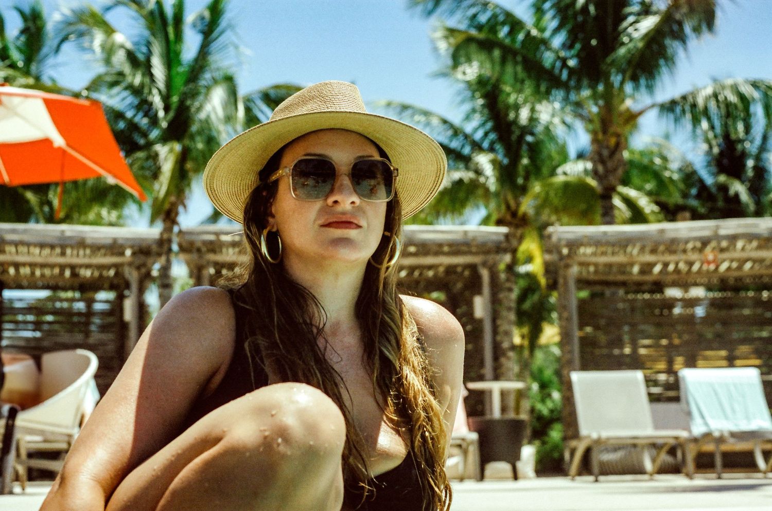 Someone in a sunhat and sunglasses lounges at a tropical resort with palm trees and beach chairs in the background.